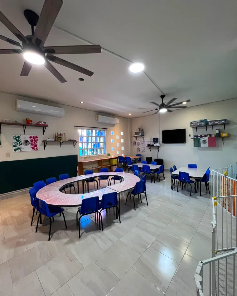Salón de kínder con mesas bajas agrupadas y sillas azules.